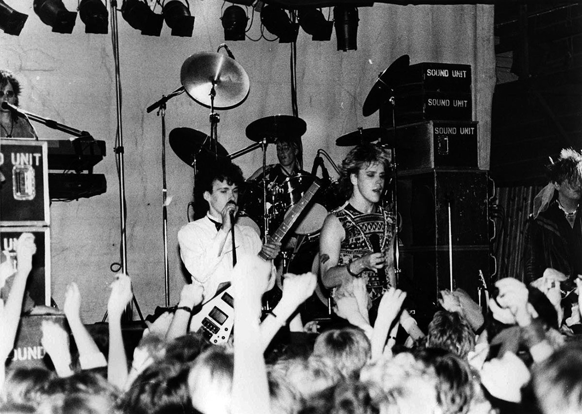 Mustavalkoisessa kuvassa taustalla Dingo-yhtye lavalla. Etualalla nuoria faneja heiluttamassa käsiään ilmassa.