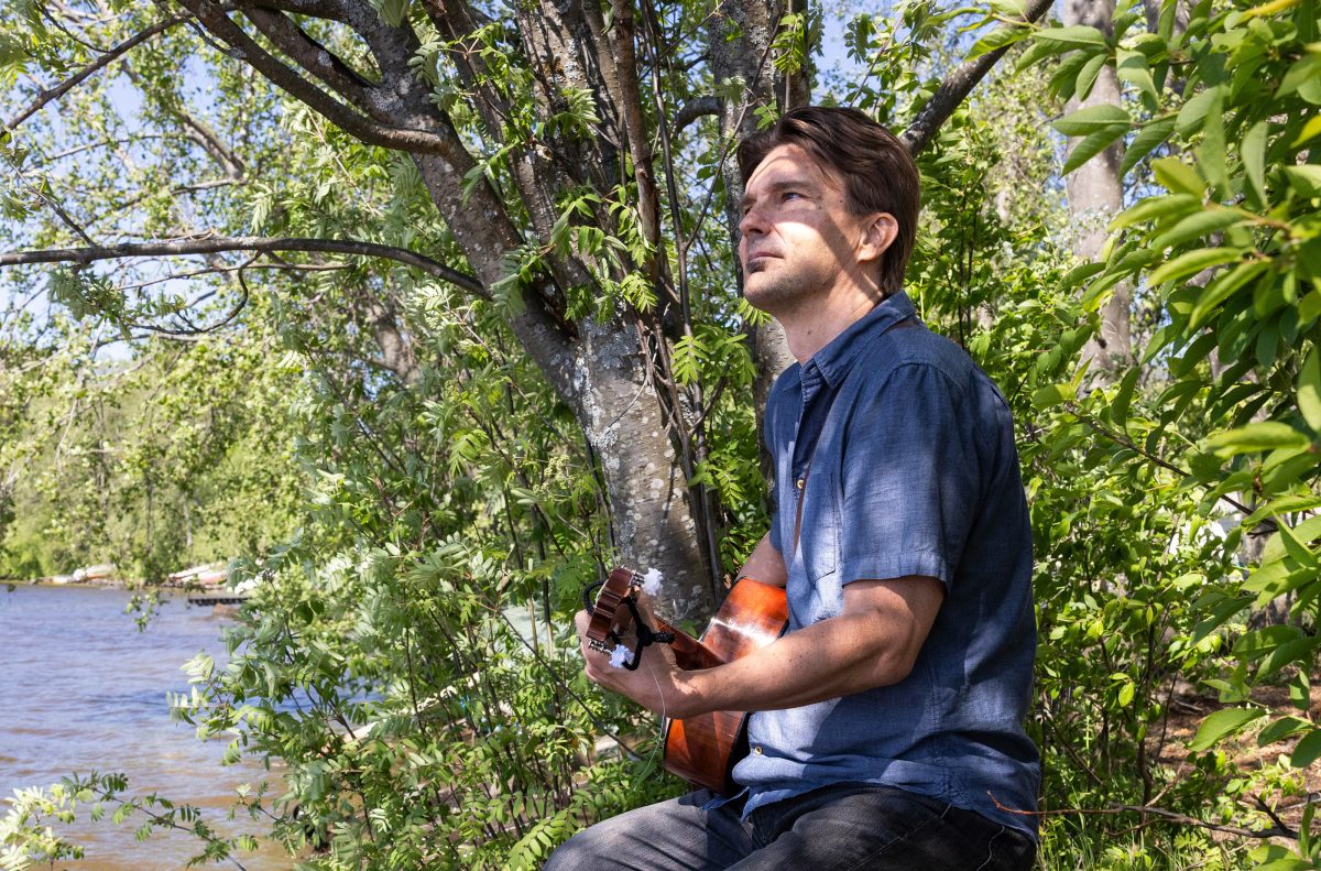 Kirjailija J. P. Laitinen istuu lehtivän puun edessä sivuittain katsojaan päin sinisessä paidassa ja soittaa nylonkielistä kitaraa.
