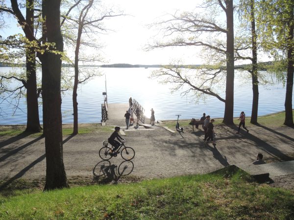 Aurinkoinen taivas ja sininen järvi. Järven rannassa on laituri, jolla seisoo perhe. Etualalla pyöräilijä ajamassa.