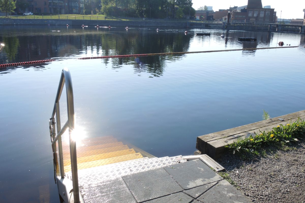 Mältinrannan uimapaikka aamulla. Etualalla metalliset portaat veteen. Taempana Tammerkosken vastaranta, jonka puut heijastuvat vedestä.