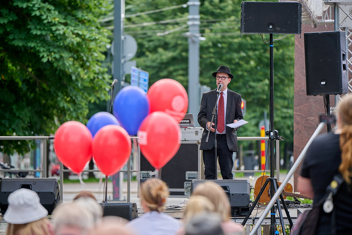 Simo Frangén puku päällä ja lierihattu päässä puhumassa Puistofiestan lavalla. Etualalla punaisia ja sinisiä ilmapalloja.