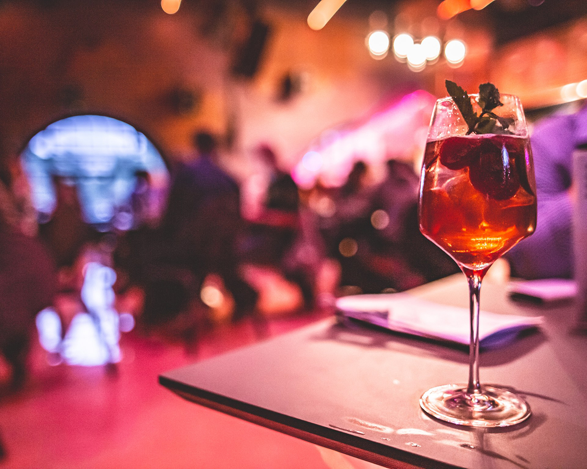 Kuva G Livelabin baaritiskiltä. Etualalla korkeajalkaisessa lasissa punaista juomaa ja juomassa kelluvia mansikoita. Taustalla violetinsävyistä valoa ja sumeasti näkyviä ihmisiä.