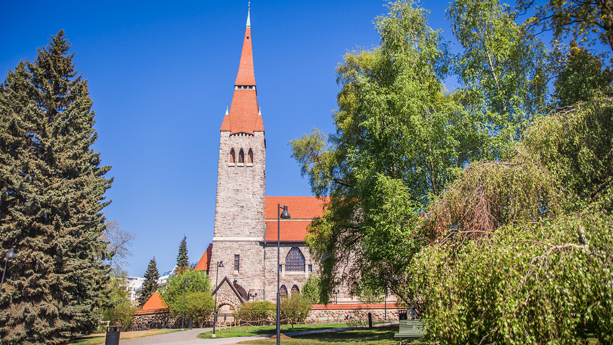 Tampereen Tuomiokirkko kesällä sinisen taivaan alla. Kirkossa on korkea harmaakivinen torni ja punaiset tiilikatot. Puistossa kirkon edessä kasvaa koivuja.